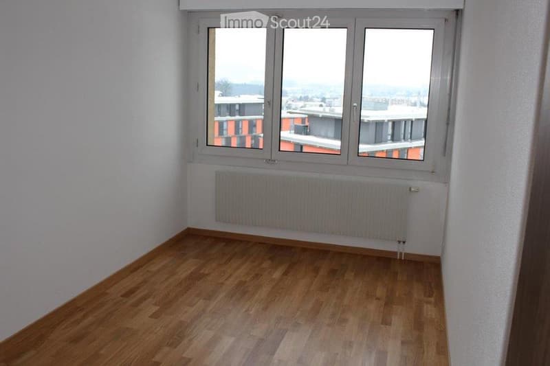 Empty room with wooden flooring, white walls, large window offering a cityscape view, and a radiator below the window.