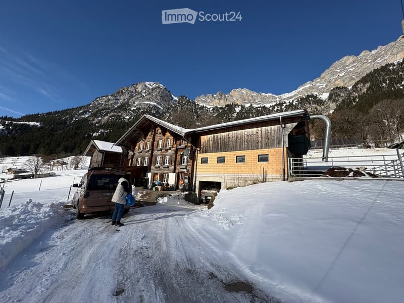 A wooden chalet-style building with a garage, situated in a snowy mountain landscape with snow-covered peaks in the background.