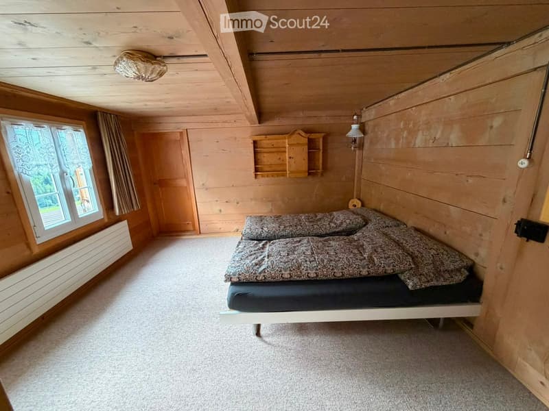 Cozy wooden bedroom with a single bed, patterned bedding, built-in wooden shelves, and a window with curtains.