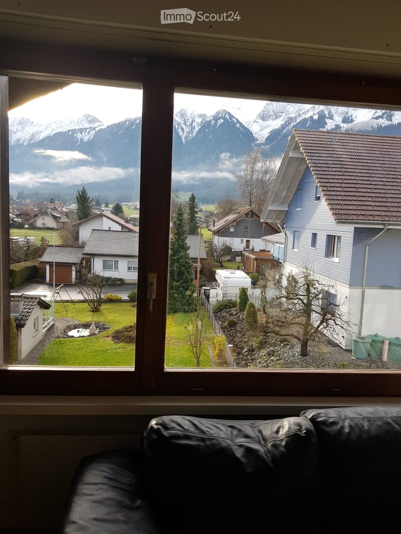 A living room with a large window offering a scenic view of a town and snow-capped mountains. The room features a black leather sofa in the foreground.