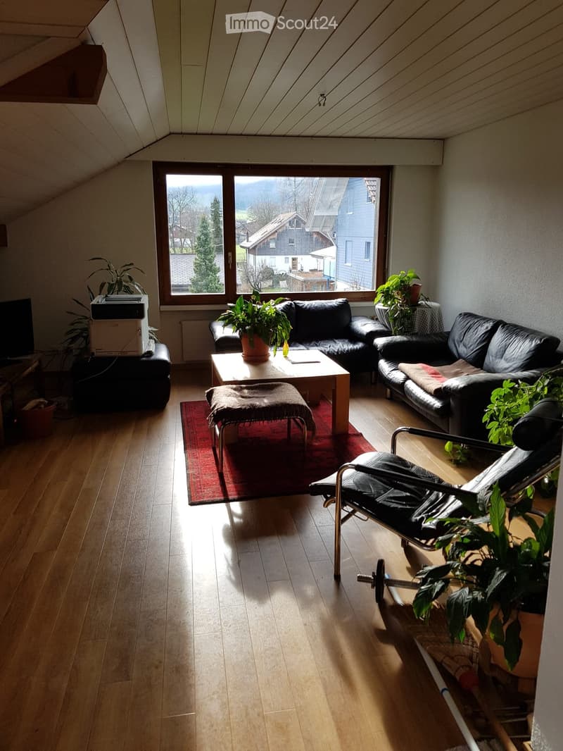 Bright living room with wooden flooring, large window offering a scenic view, black leather sofa, coffee table, chair, and plants.