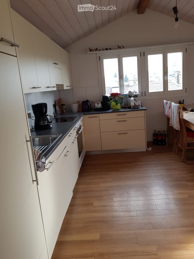 Modern kitchen with light wooden flooring, white cabinets, and various appliances including a coffee maker, oven, and sink. There is a small dining area with a table and chair.