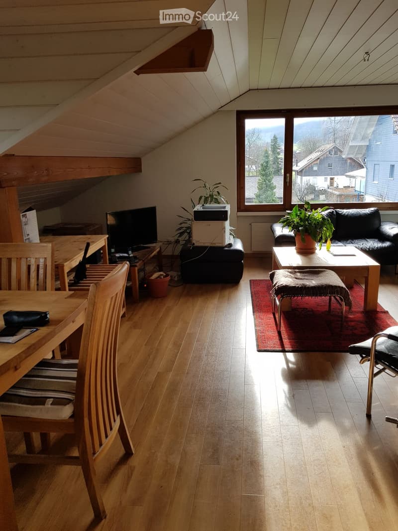Bright living room with wooden floors, large window offering a view, sofa, coffee table, small table, and dining area with chairs.