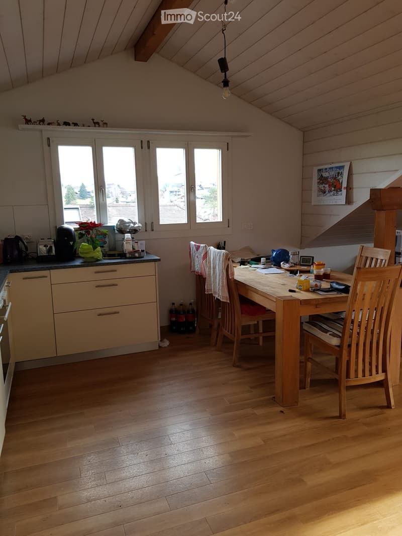 Bright kitchen with wooden flooring, white cabinets, and a large window. Includes a wooden dining table with chairs, a coffee maker, and various kitchen items on the counter.
