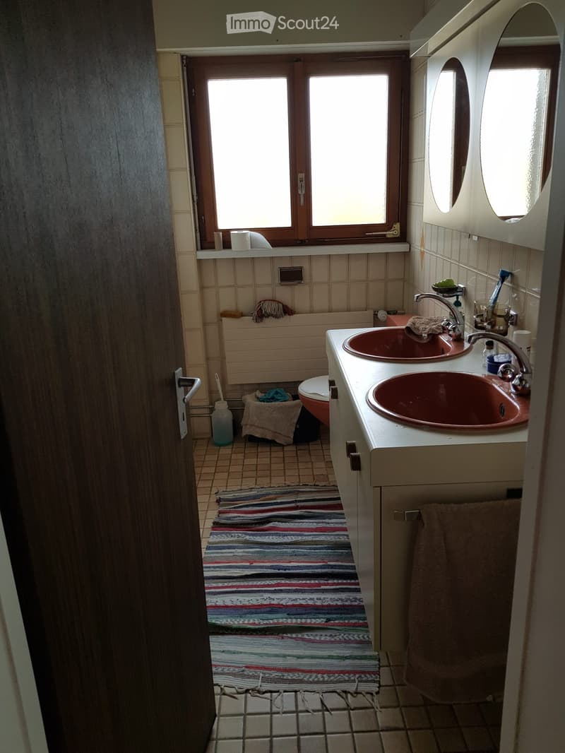 Bathroom with double sink, mirror, toilet, and small window. Includes a striped rug on the floor and various toiletries on the sink counter.