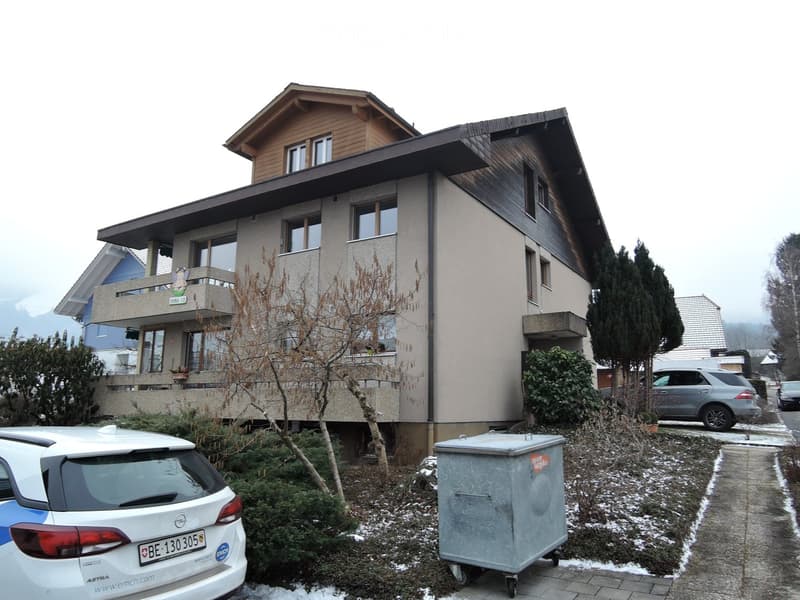 Two-story house with a wooden roof, multiple windows, balconies, and surrounding greenery.