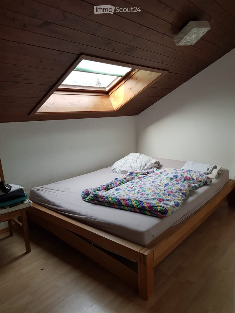 Cozy attic bedroom with wooden slanted ceilings, skylight window, single bed with colorful quilt and pillows, wooden floor, and a small wooden chair with folded clothes.