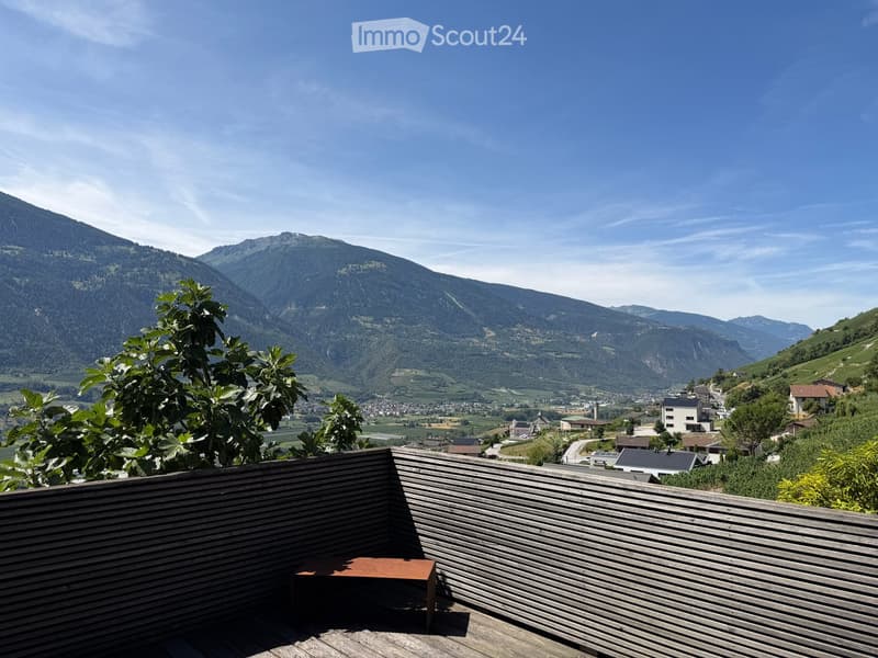 Wooden balcony with a view of a town and mountains, featuring a small table and lush greenery.