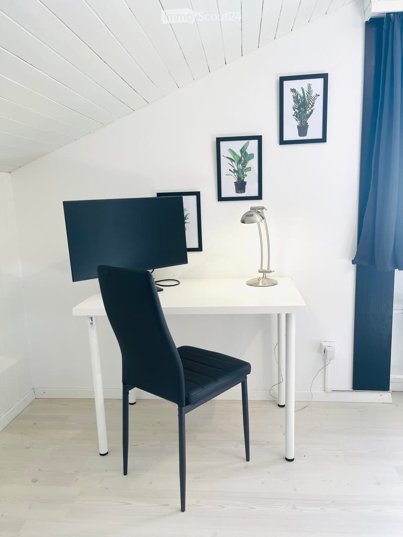 A modern, minimalist workspace with a white desk, black chair, sleek lamp, and large screen, complemented by framed artwork and blue curtains.