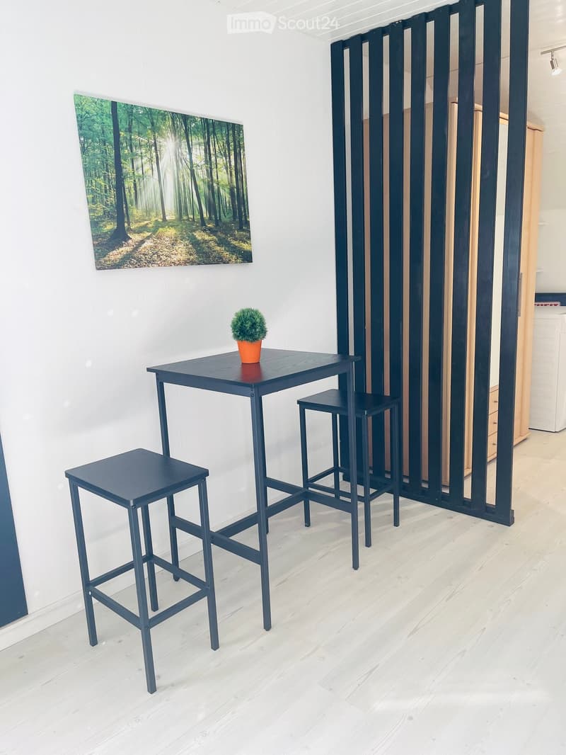 A small, modern room with a dark blue table, two matching stools, and a small potted plant on the table. A large black screen or partition is visible in the background, along with a large forest-themed painting on the wall.