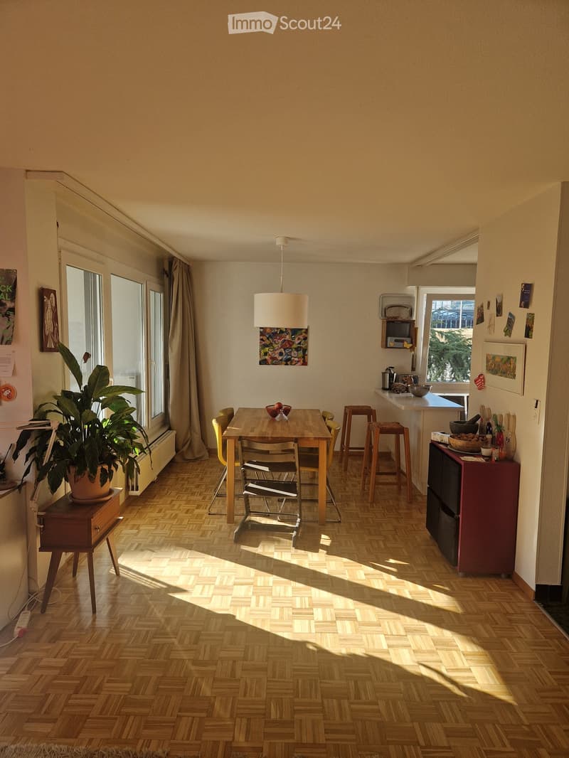Bright, open kitchen-dining area with wooden herringbone flooring, large windows, a dining table with chairs, stools, a refrigerator, and a potted plant near the window.