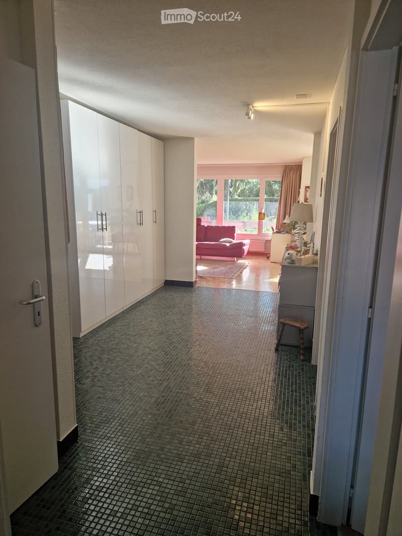 A narrow hallway with tiled flooring, white walls, and built-in white cabinets on one side. The hallway leads to a brightly lit living area with a red sofa, a small table, and a lamp.