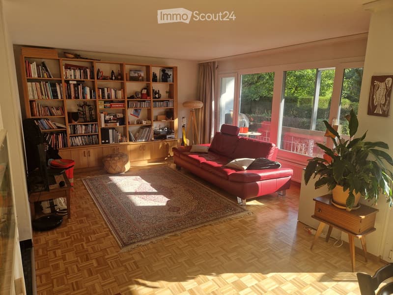 Bright living room with wooden flooring, large windows, red leather sofa, bookshelf, small table, and potted plant.
