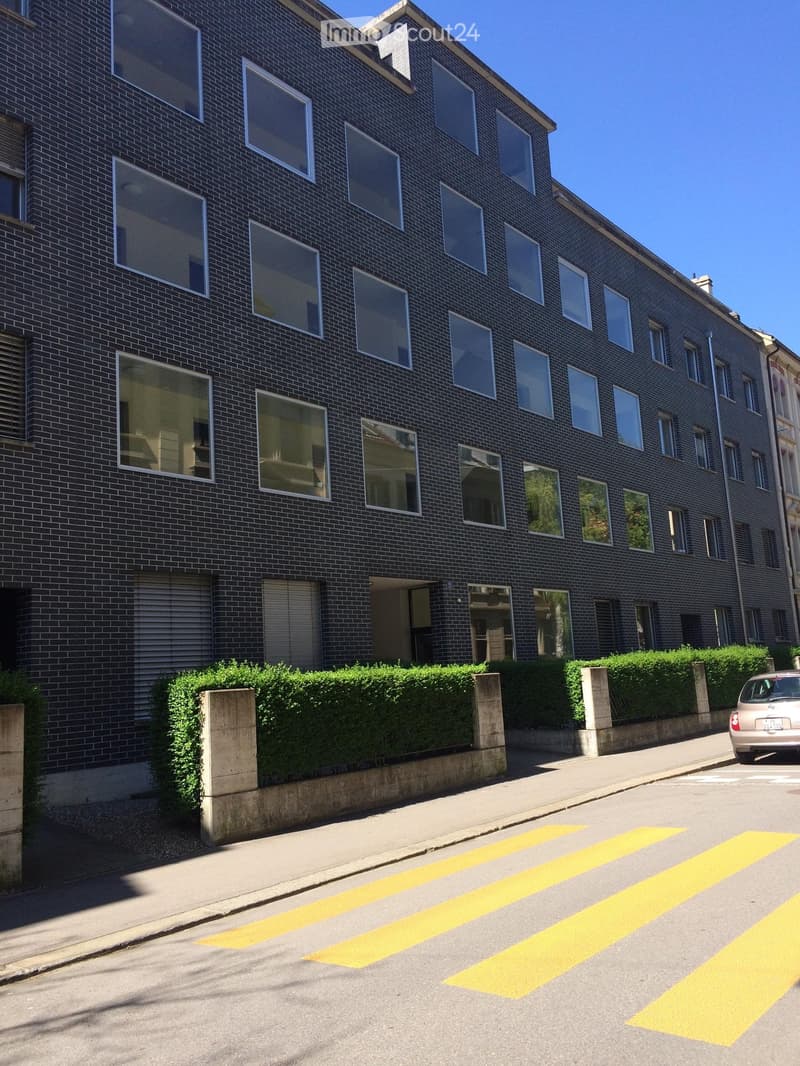 Multi-story building with dark brick facade and large windows, featuring a modern design and greenery along the sidewalk.