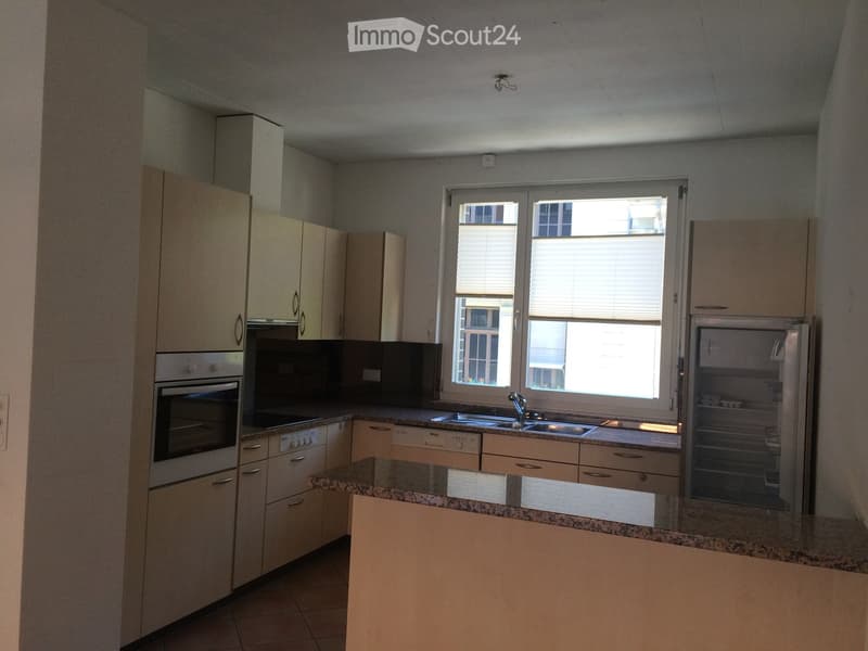 Modern kitchen with light-colored cabinets, granite countertops, stainless steel appliances including an oven, sink, and refrigerator, and a large window with blinds.