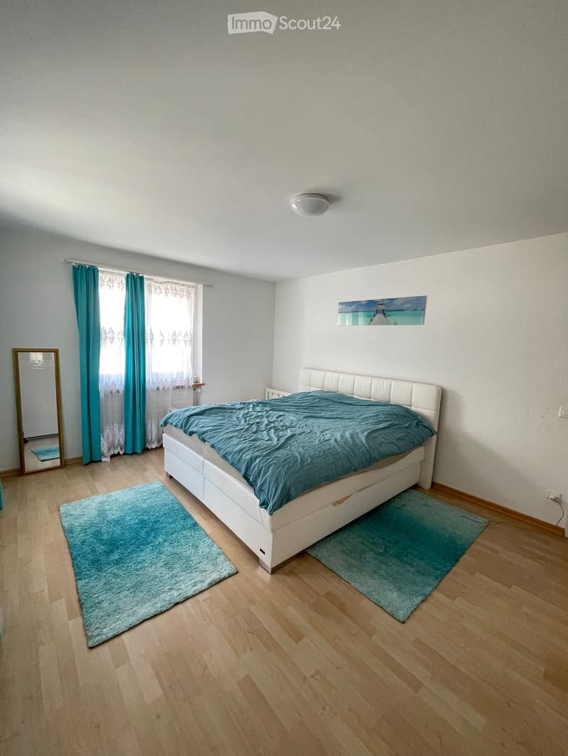 Bright bedroom with wooden flooring, white bed with teal bedding, two teal rugs, large window with teal curtains, and a mirror on the wall.