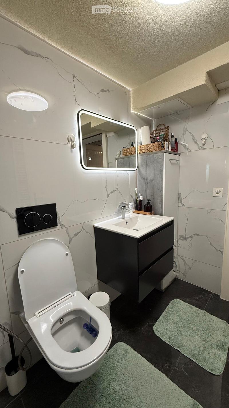 Modern bathroom with white marble walls, a toilet, a sink with a black cabinet, a large mirror, and two green rugs on the dark floor.