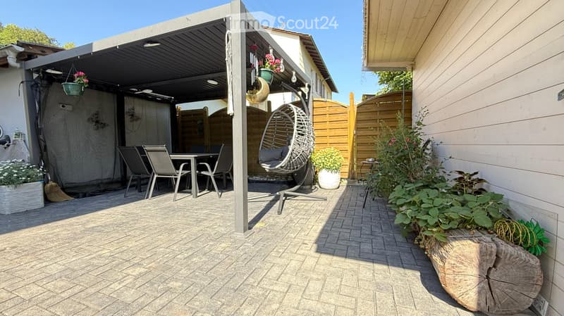 A covered patio area with a dining table and chairs, a hanging egg chair, potted plants, and a wooden fence.
