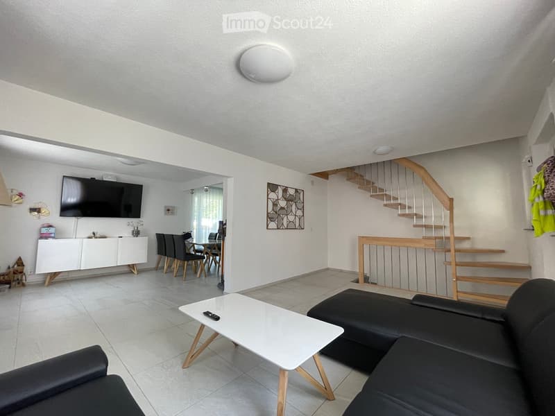 Modern living room with a black sofa, white coffee table, flat-screen TV, dining area with chairs, and a staircase leading to an upper level.
