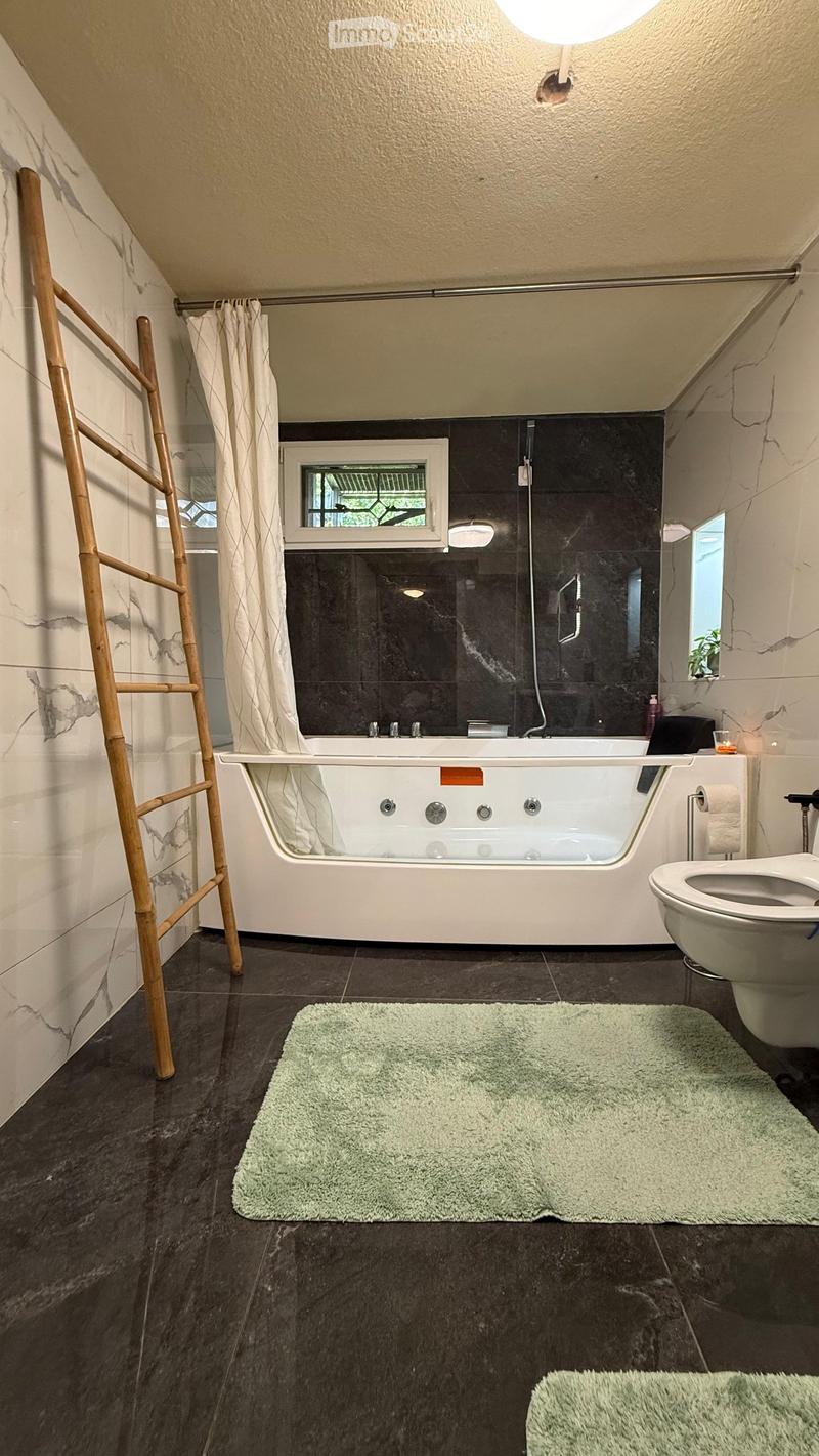 Modern bathroom with a large bathtub, toilet, shower area with dark tiles, and a window. Includes a bamboo towel rack and green bathroom rugs.