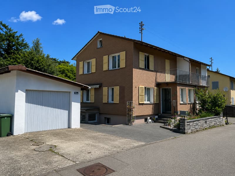 Two-story house with brown brick facade, white garage, and balcony on the upper floor. The house features multiple windows with shutters and a small garden area in front.