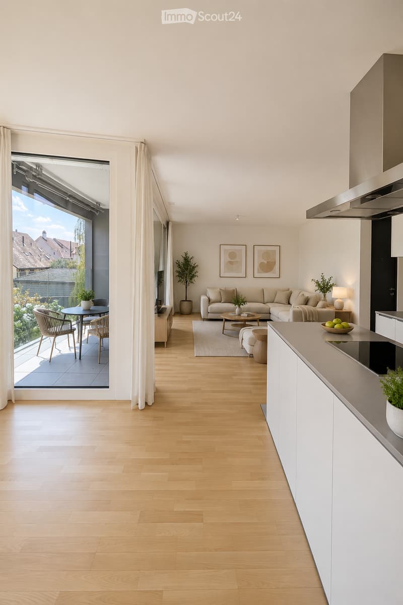 Modern open-plan living and dining area with light wood flooring, white walls, and large windows. Features a grey sectional sofa, coffee table, dining table with chairs, kitchen counter, and decorative plants.