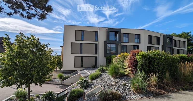 Modern two-story house with large windows, grey facade, and landscaped garden with shrubs and trees.