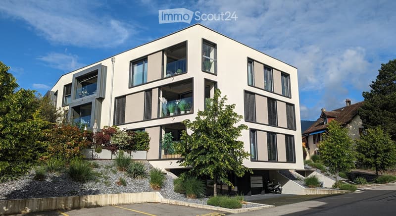 Modern multi-story building with large windows and balconies, surrounded by greenery and a paved parking area.