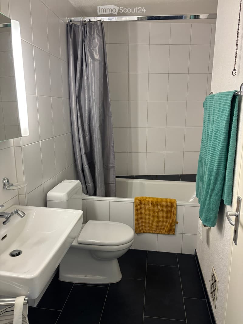 Modern bathroom with white tiles, a sink, toilet, bathtub, and towels hanging on the wall.