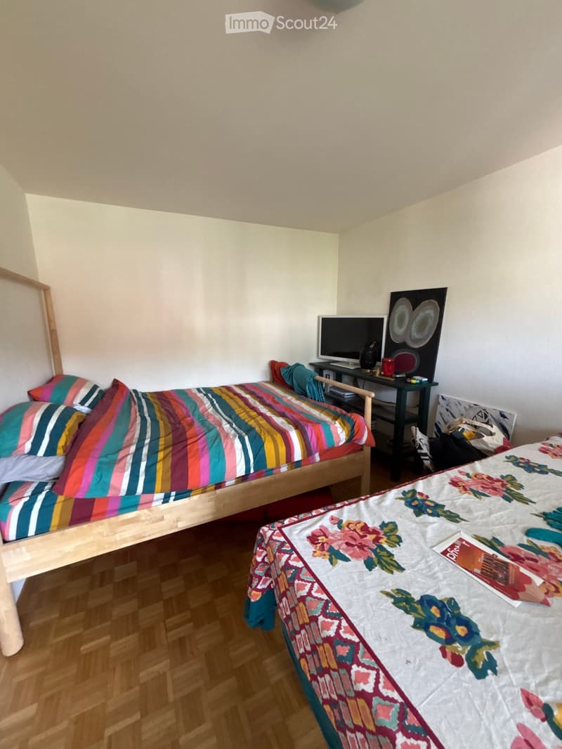 A small bedroom with two single beds, one with a colorful striped duvet and the other with a floral duvet. A small table with a screen and some items is placed between the beds.