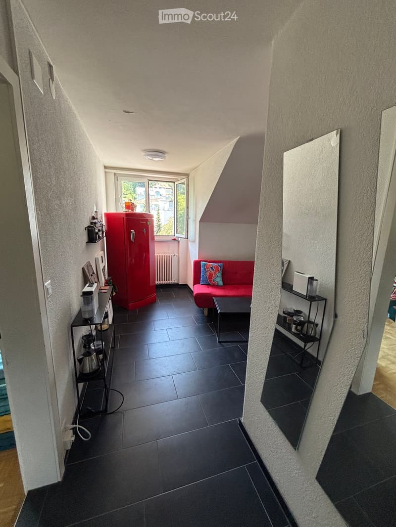 A small, modern living room with a red sofa, coffee table, and a red refrigerator. The room features dark tiled flooring and a view through a large window.