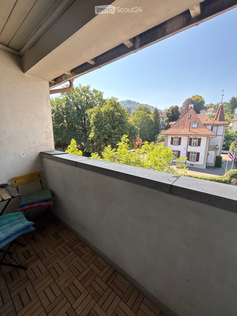 Balcony with wooden floor, a small table, and a chair with cushions, overlooking a scenic view of trees and a historic building.