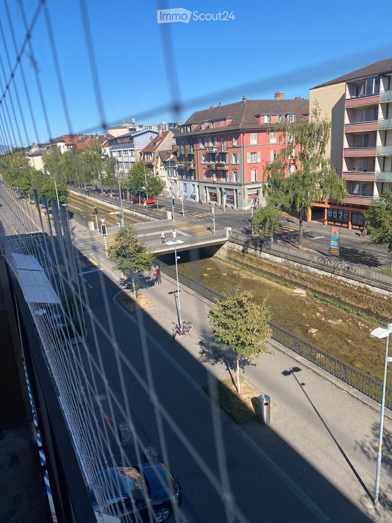 A view of a city street with buildings, traffic, and a canal running alongside. The scene includes a bridge, pedestrians, and a parked car visible through the foreground.