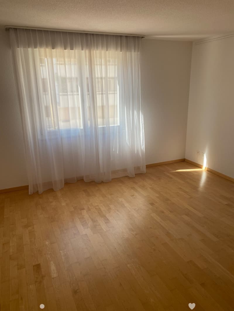 Empty room with wooden flooring and white walls, featuring a large window with sheer curtains allowing natural light to enter.