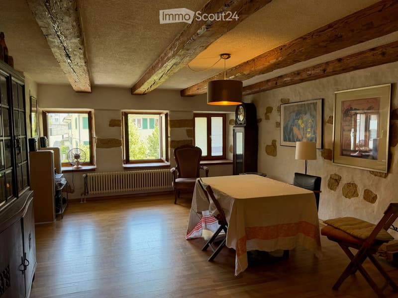 Dining area with wooden floor, large windows, table with chairs, fan, cabinet, lamp, and artwork on the walls.