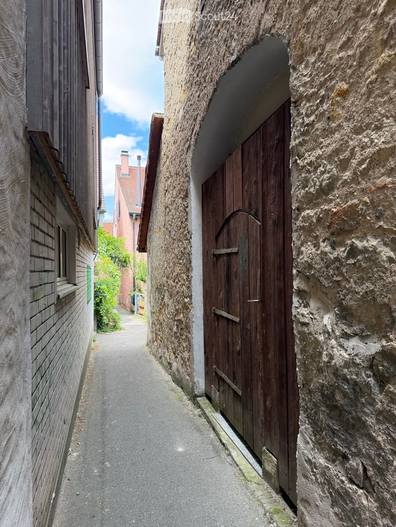 Narrow alleyway between two buildings, one with a wooden door and stone walls, the other with brick walls and a small window. The alley is paved and leads to a small garden area with greenery.
