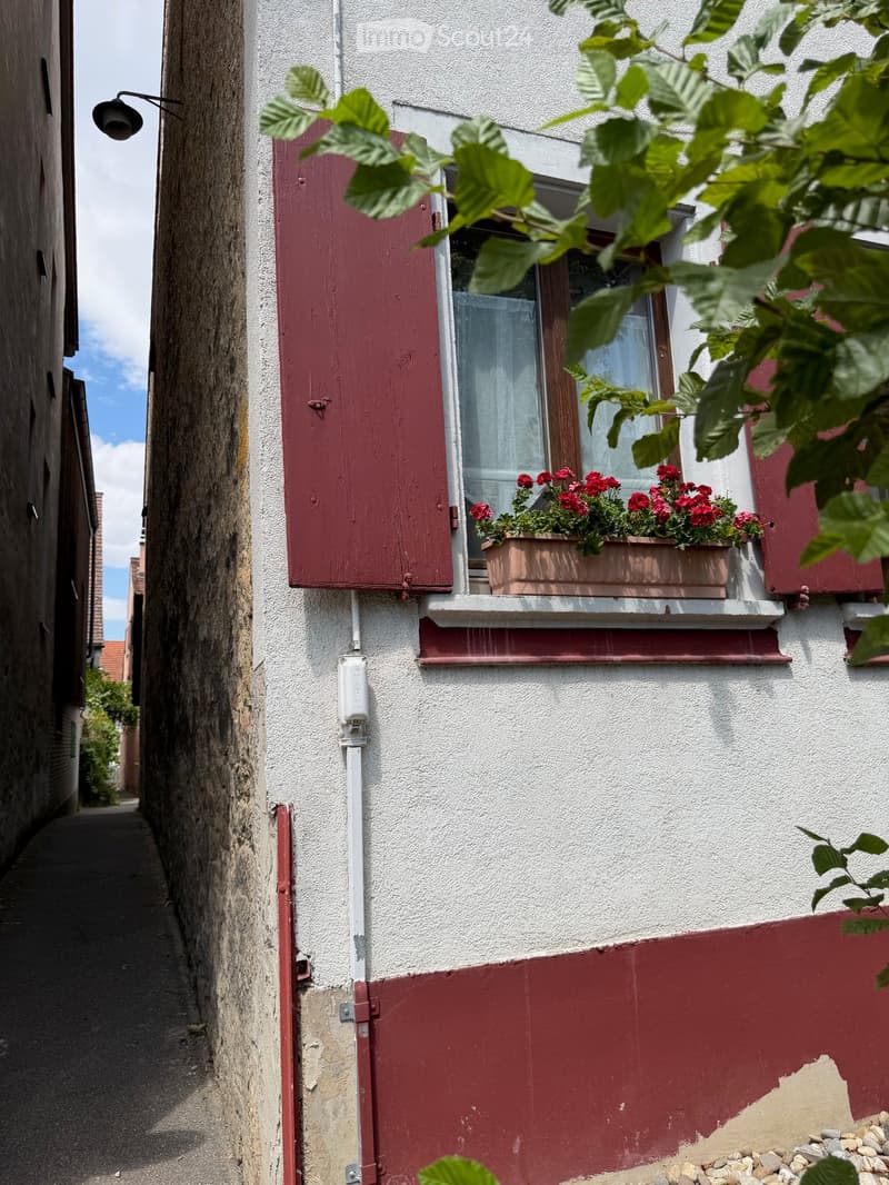 A narrow alleyway with a white building featuring red shutters and a window box with red flowers. The building has a mix of white and red exterior walls.