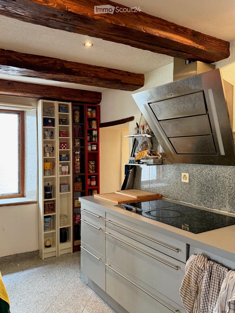 Modern kitchen with grey cabinets, induction stove, oven, and wooden cutting board on the island. Shelves hold various kitchen items, including coffee maker and blender. Exposed wooden beams on the ceiling.