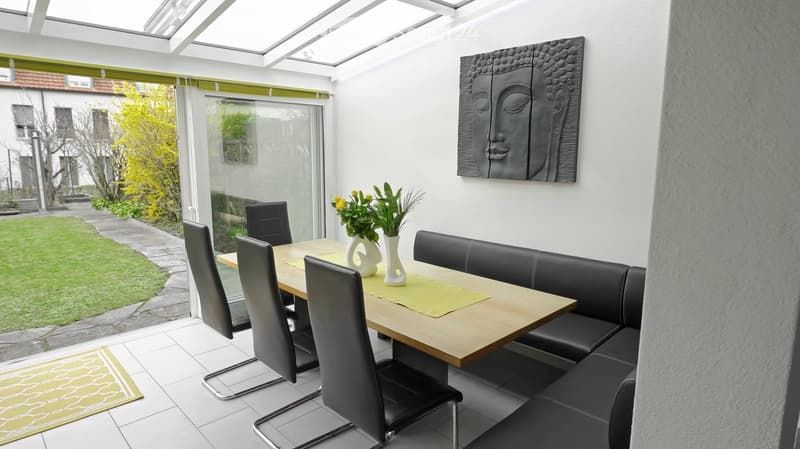 Bright dining area with a wooden table, black leather sofa, chairs, vases, and a large wall painting, overlooking a garden.
