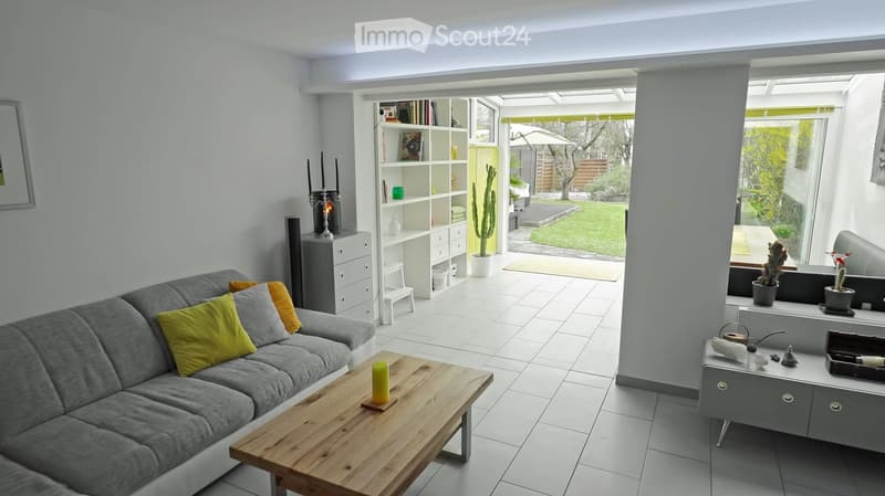 Modern living room with grey sofa, wooden coffee table, and white tiled floor. Features a bookshelf, small cabinet, and a view of a garden through large windows.