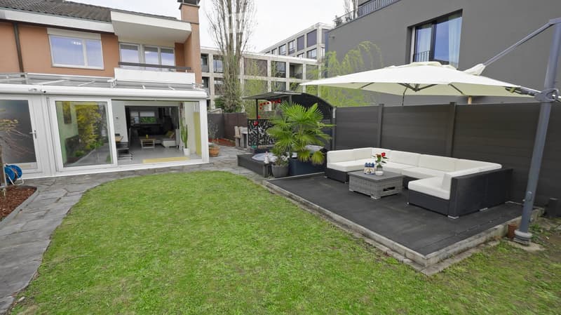 Well-maintained garden with a lawn, patio area, outdoor seating, and a large umbrella. Adjacent to a modern house with large windows and a balcony.