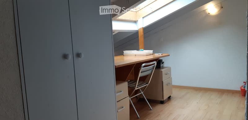 A small, well-lit room with wooden flooring, a desk, a chair, and a cabinet. The room features a skylight and a built-in storage unit.