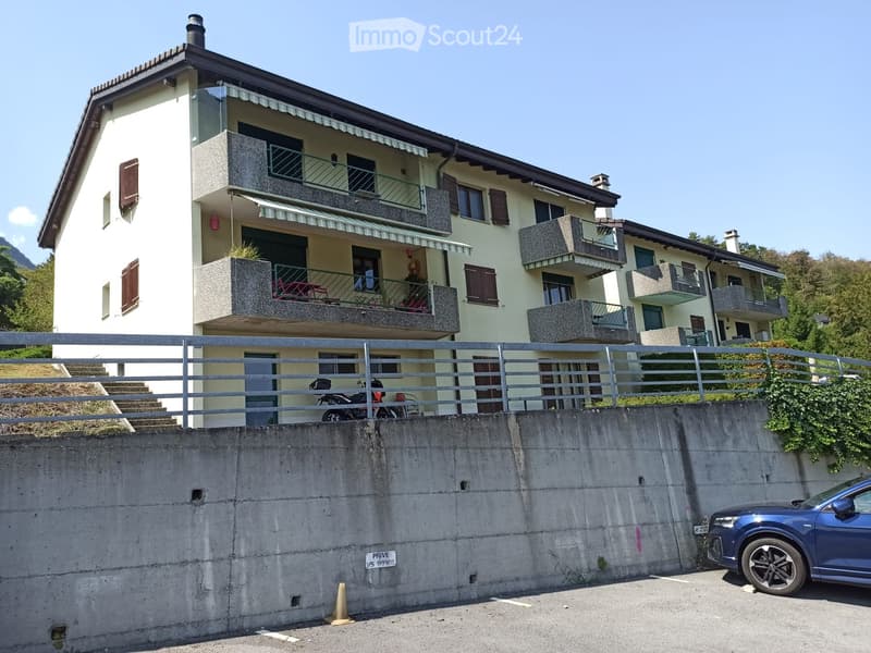Two-story house with balconies, multiple windows, and a staircase leading to the entrance. A car is parked nearby, and there is a concrete wall in the foreground.