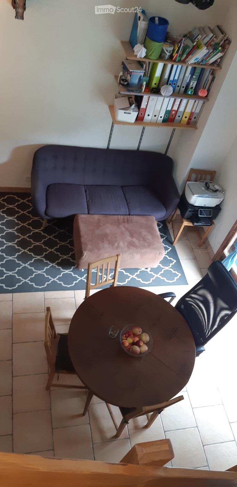 A small, cozy living room with a sofa, a round wooden table with chairs, a bookshelf filled with books and binders, and a printer on a small table. The room features a patterned rug and a cushioned seat.