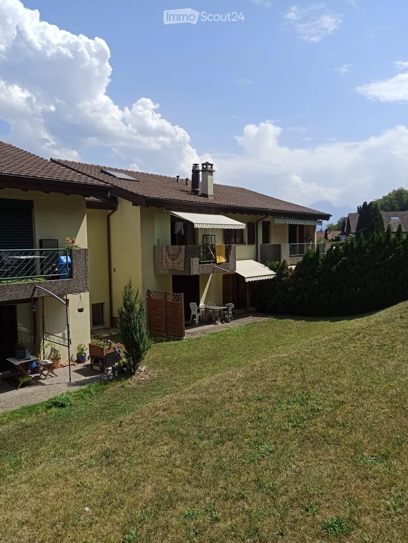 A two-story house with a tiled roof, multiple balconies, and a well-maintained lawn. Outdoor seating area with a table and chairs, small garden beds, and a wooden fence.