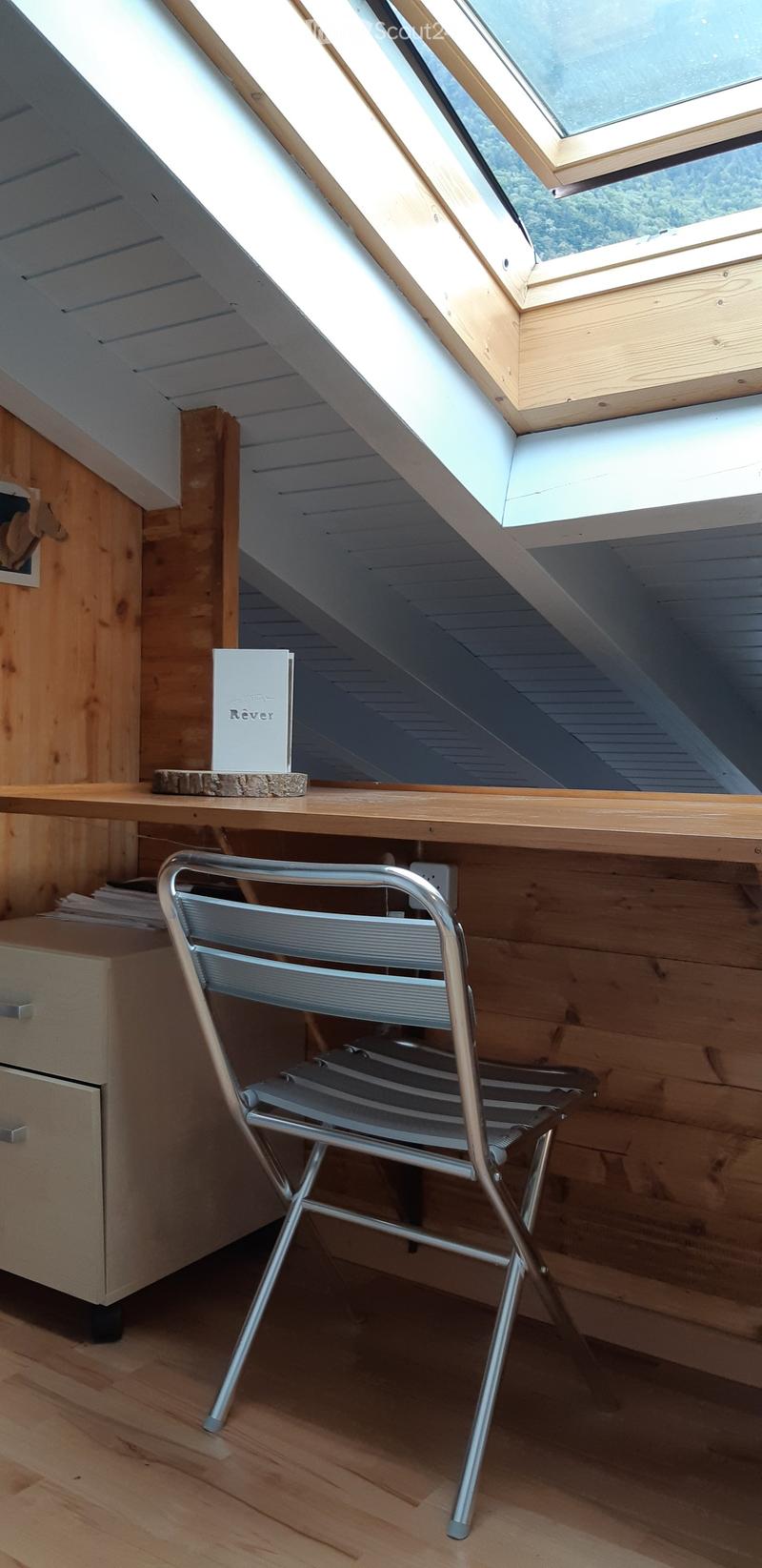 Loft-style room with wooden walls and ceiling, featuring a slanted roof with skylights, a wooden table with a book, a metal folding chair, and a small cabinet.