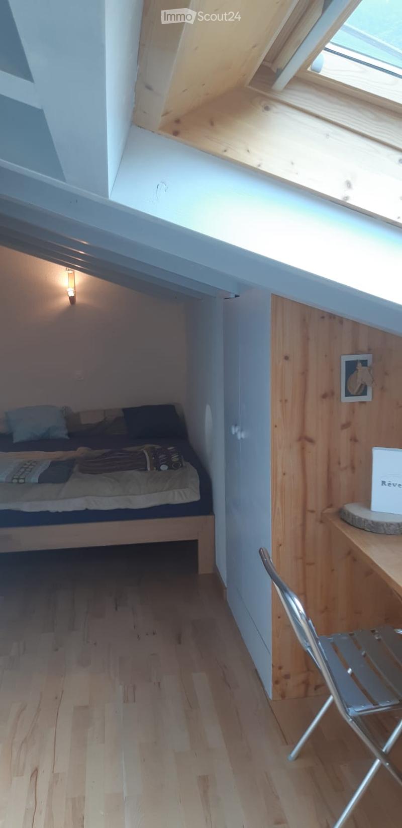 Cozy attic bedroom with wooden floors, slanted ceilings, and a skylight. Features a small bed, a wooden table with a chair, and a picture frame on the wall.