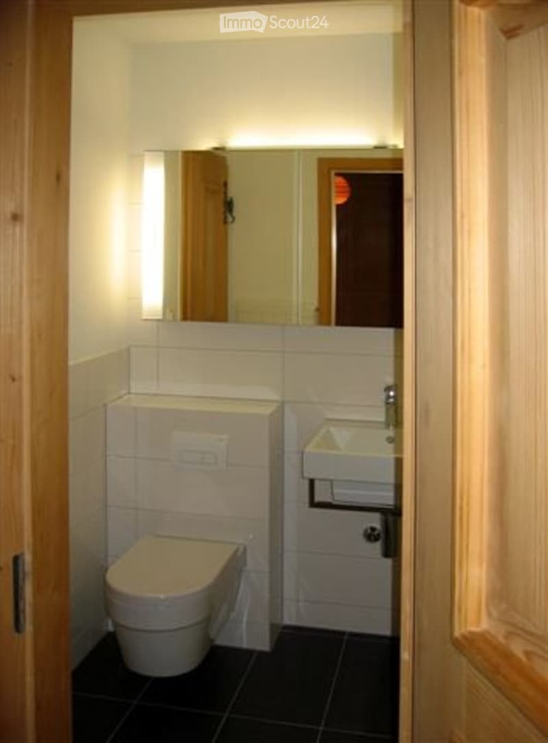 Compact bathroom with white tiles, a toilet, a sink, and a mirror above it. The walls are tiled, and the floor is dark.