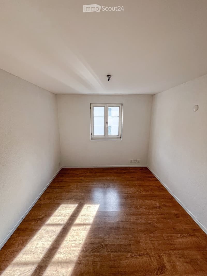 Empty room with light-colored walls, wooden flooring, and a single window allowing natural light to enter.