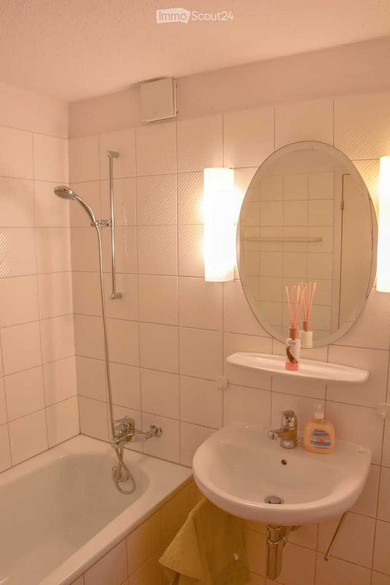 Modern bathroom with a bathtub, sink, mirror, and showerhead. Includes a soap dispenser and a towel on the floor.